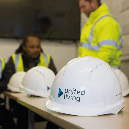 United Living hard hats placed on a table with construction workers sat in the background