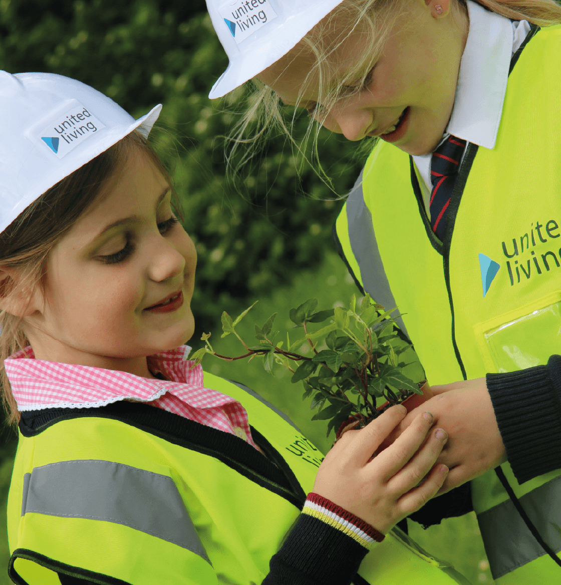 Two children wearing Unitet Living hard hats and high vis jackets holding a plant